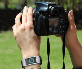 Color close-up photo of two hands holding a digital single-lens reflex camera; on the left wrist is a band with a digital display