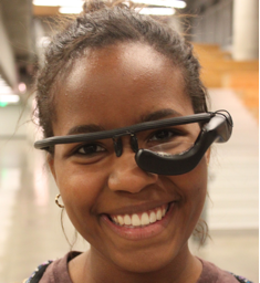 Close-up photo of young smiling African-American woman wearing black eyeglass frame fitted with a sensing device below her left eye